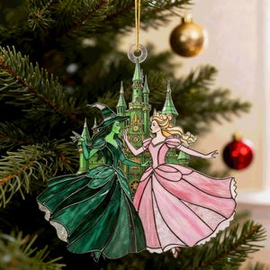 Puede incluir: Adorno navideño con una bruja verde y una princesa rosa frente a un castillo verde. El adorno cuelga de un hilo dorado. El fondo incluye un árbol de Navidad y otros adornos.