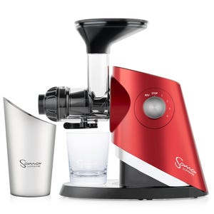 May include: A red Sana Supreme juicer with a black and silver base. The juicer has a clear juice spout and a black feeding tube. The juicer has a dial with the words "Rev Stop" and numbers 1 through 4. The juicer is sitting on a white surface. There is a silver cup with the Sana Supreme logo and a clear glass on the surface.