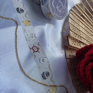 May include: A gold chain, decorative washi tape rolls, and a decorative fan are displayed on a white surface. The washi tape features a repeating pattern of cartoon characters. One roll has the text "Deepspace Washi". A red, textured item is also visible.