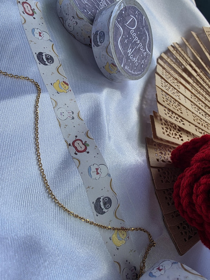May include: A gold chain, decorative washi tape rolls, and a decorative fan are displayed on a white surface. The washi tape features a repeating pattern of cartoon characters. One roll has the text "Deepspace Washi". A red, textured item is also visible.