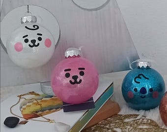 Puede incluir: Una colección de adornos navideños redondos y brillantes. Uno es blanco con una cara de dibujos animados y mejillas rosadas, otro es rosa y un tercero es azul. Cada adorno tiene una tapa plateada y un lazo para colgar.