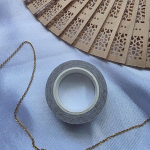 May include: A roll of gray patterned washi tape with a white core sits next to a gold chain and a tan, intricately carved wooden fan. The fan has a delicate, openwork design. The items are on a light blue fabric.