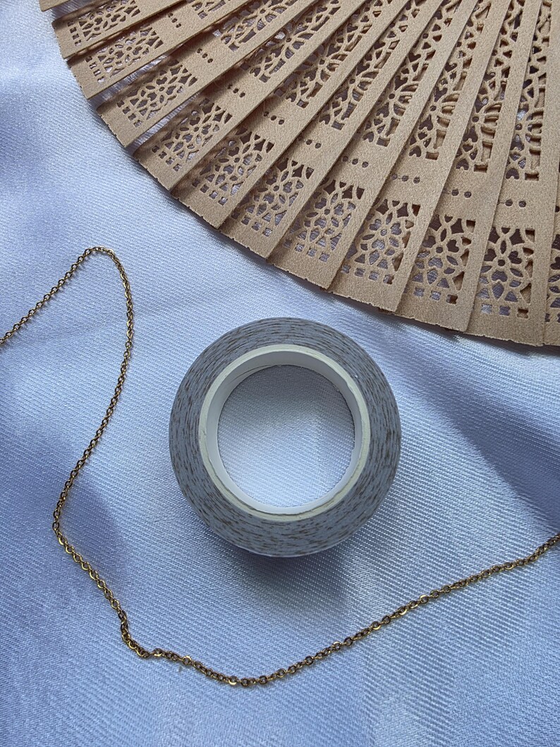 May include: A roll of gray patterned washi tape with a white core sits next to a gold chain and a tan, intricately carved wooden fan. The fan has a delicate, openwork design. The items are on a light blue fabric.