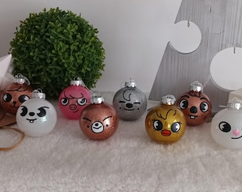 Puede incluir: Una colección de adornos navideños brillantes con caras de dibujos animados. Los adornos son de varios colores, incluyendo marrón, rosa, plata, oro y blanco. Cada adorno tiene una tapa plateada para colgar. Un arbusto verde y un árbol decorativo están en el fondo.