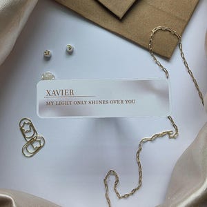 Puede incluir: Un collar de cadena dorada con un colgante rectangular que dice "XAVIER MY LIGHT ONLY SHINES OVER YOU". La imagen tambi&eacute;n presenta pendientes dorados en forma de estrella, clips y un sobre peque&ntilde;o, todo sobre una superficie blanca.