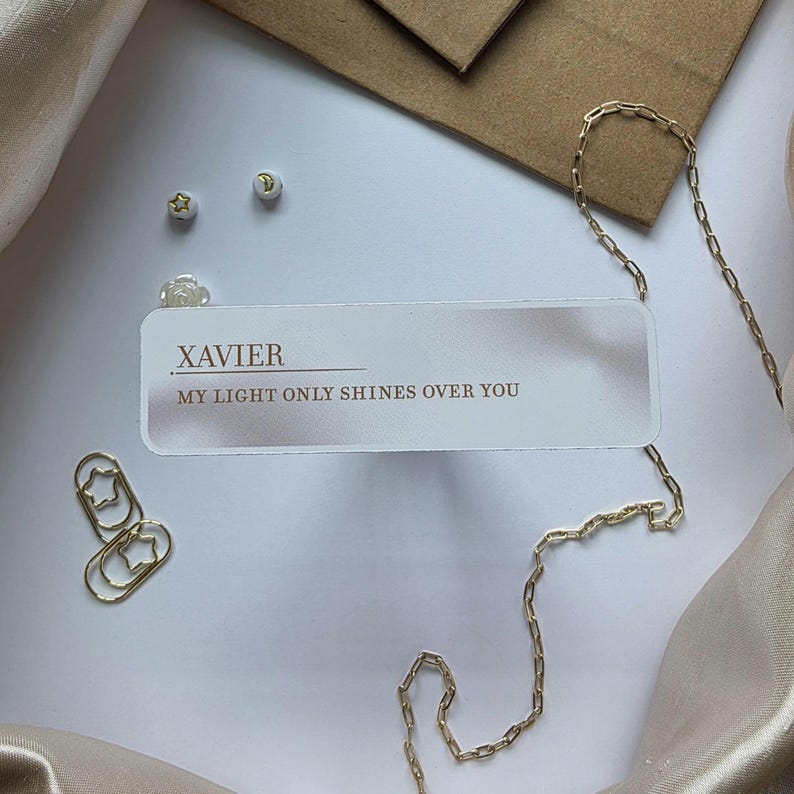 Puede incluir: Un collar de cadena dorada con un colgante rectangular que dice "XAVIER MY LIGHT ONLY SHINES OVER YOU". La imagen tambi&eacute;n presenta pendientes dorados en forma de estrella, clips y un sobre peque&ntilde;o, todo sobre una superficie blanca.