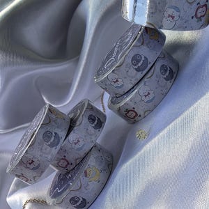 May include: Several rolls of decorative washi tape with a light gray background and a repeating pattern of cartoon characters in various colors. The tape rolls are stacked and arranged on a white satin surface.