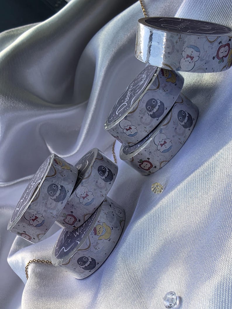 May include: Several rolls of decorative washi tape with a light gray background and a repeating pattern of cartoon characters in various colors. The tape rolls are stacked and arranged on a white satin surface.