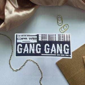 May include: A white and black sticker with the text "GANG GANG" in a glitch font. The sticker also has the text "DPR WE" and a barcode design. Gold star-shaped paper clips and a gold chain are also visible.