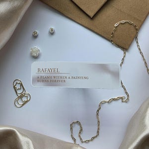 Puede incluir: Una vista cenital presenta joyas y art&iacute;culos de papeler&iacute;a. Una cadena dorada, clips y peque&ntilde;os pendientes con forma de estrella, luna y flor se distribuyen alrededor de una tarjeta con el texto "RAFAYEL A FLAME WITHIN A PAINTING BURNS FOREVER."