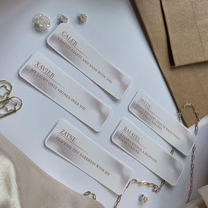 May include: Five rectangular, frosted acrylic name tags with gold text and quotes. The names are Caleb, Xavier, Zayne, Sylus, and Rafayel. The tags are arranged on a white surface with gold jewelry and a satin fabric.