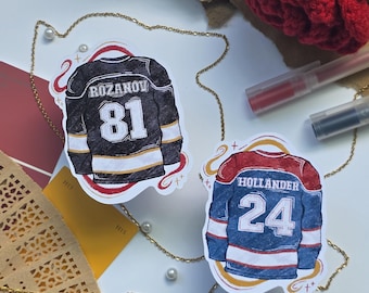 Puede incluir: Dos pegatinas de camisetas de hockey, una negra con "Rozanov 81" y otra azul con "Hollander 24". También se ven una rosa roja de ganchillo y una cadena dorada.