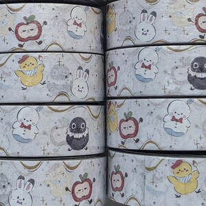 May include: Rolls of decorative washi tape featuring repeating patterns of cartoon characters, including apples, bunnies, chicks, and penguins. The tape has a light gray background with gold accents and small cross symbols.