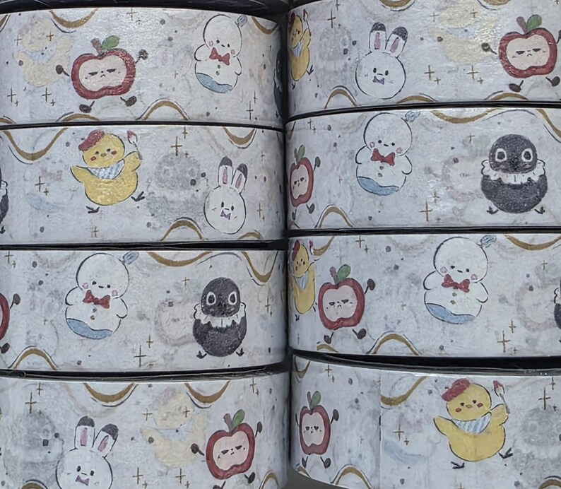 May include: Rolls of decorative washi tape featuring repeating patterns of cartoon characters, including apples, bunnies, chicks, and penguins. The tape has a light gray background with gold accents and small cross symbols.