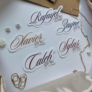 May include: White stickers with names in elegant cursive script, including "Rafael," "Xavier," "Zayne," "Sylus," and "Caleb." Each sticker has the word "girlie" below the name. Gold chain and star-shaped paper clips are also visible.