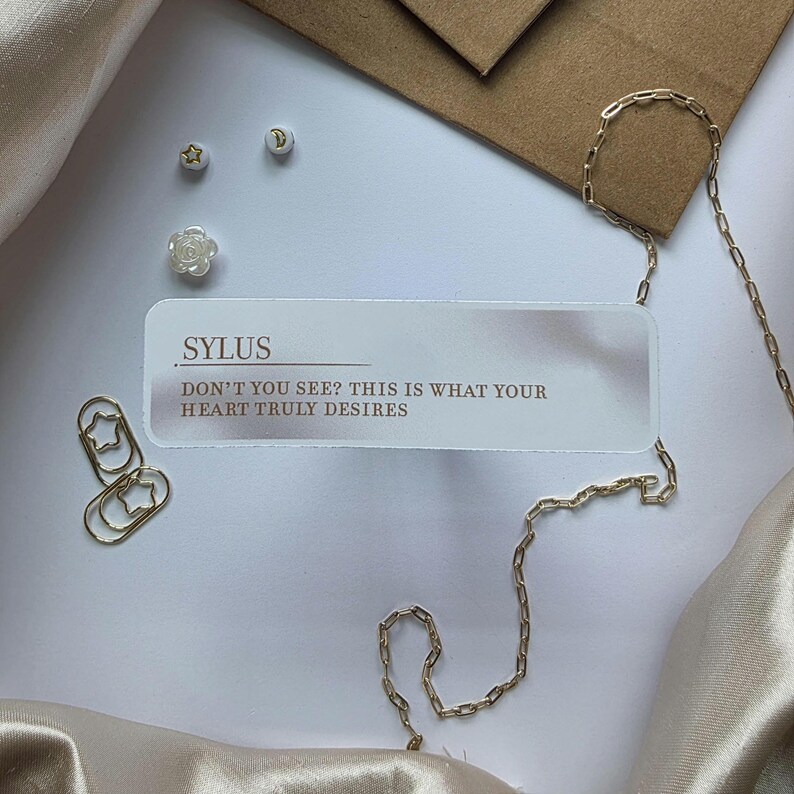 Puede incluir: Composici&oacute;n plana con cadena dorada, clips y peque&ntilde;os elementos decorativos. Una tarjeta con la palabra "SYLUS" y el texto "DON'T YOU SEE? THIS IS WHAT YOUR HEART TRULY DESIRES" est&aacute; presente. Fondo con sobre marr&oacute;n y tela drapeada.