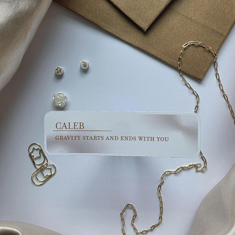 Puede incluir: Composici&oacute;n plana con un collar de cadena dorada, un clip y peque&ntilde;os pendientes. Una tarjeta con el nombre "CALEB" y la frase "GRAVITY STARTS AND ENDS WITH YOU" est&aacute; presente. El fondo incluye un sobre marr&oacute;n y tela color crema.