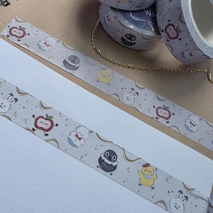 May include: Several rolls of decorative washi tape with a light gray background and repeating patterns of cartoon characters, including apples, bunnies, and chicks. The tape is unrolled on a white surface, revealing the design. The text "Washi" is visible on the tape rolls.