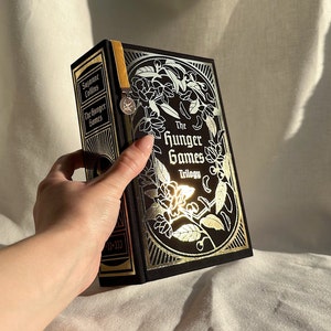 Può includere: Un libro rilegato nero con dettagli in oro. Il libro è intitolato "The Hunger Games Trilogy" di Suzanne Collins. La copertina presenta un design floreale argentato.