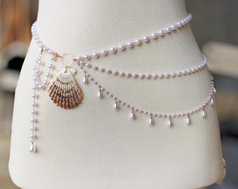May include: A multi-strand waist chain featuring white faux pearls and a central natural seashell pendant. The design includes three draped pearl strands, a dangling pearl accent, and small teardrop-shaped pearl charms hanging from the lower chain.