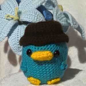 May include: A crocheted plush toy of a blue platypus wearing a brown hat. The platypus has a yellow beak and black eyes.