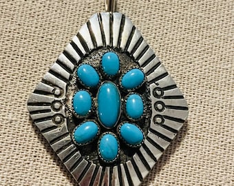 May include: A silver pendant with a geometric design and turquoise stones. The pendant has a diamond shape with a sunburst design around the edge. The turquoise stones are set in silver and are arranged in a circular pattern.