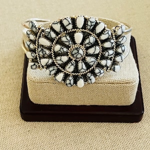 Puede incluir: Una pulsera de plata con un diseño floral central. El diseño está compuesto por piedras blancas y negras, rodeadas por una cuerda de plata retorcida. La pulsera se exhibe sobre un soporte cubierto de tela beige.