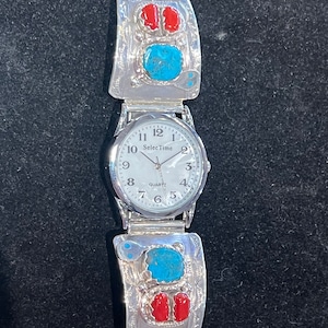 May include: A silver watch with a white face and black numbers. The watch has a silver band with turquoise and red stones.