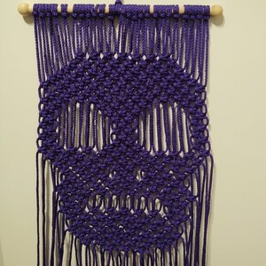 May include: A purple macrame wall hanging with a circular design. The hanging is made with knotted cords and has a wooden dowel at the top.