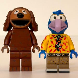 May include: Two Lego figures of Muppets characters. One is a brown dog with long ears and a wide smile. The other is a blue bird-like character with a purple beak, wearing a yellow shirt with red chili peppers and a black tie.