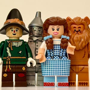May include: Four Lego figures representing characters from The Wizard of Oz. The figures include the Scarecrow in a black hat and green outfit, the Tin Man in silver, Dorothy in a blue gingham dress, and the Cowardly Lion.