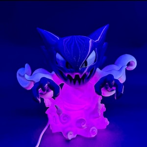 May include: A blue and white figurine of a cartoon ghost with a menacing expression and sharp teeth. The figurine has a glowing pink cloud base and is lit with a purple light. The figurine has a cable.