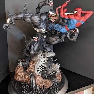 May include: A detailed statue featuring the characters Venom and Spider-Man in a dynamic battle pose. Venom is depicted in black with a white spider symbol, while Spider-Man is in his classic red and blue suit. The base has a detailed, textured design.
