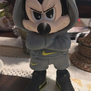 Puede incluir: Figura de Mickey Mouse con sudadera gris, pantalones cortos y zapatillas negras con detalles amarillos. La figura tiene una expresión seria. El logo de Nike es visible.