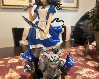 May include: A detailed figurine of Alice in Wonderland, dressed in a blue and white dress, holding a mallet and a large knife. She stands atop a creature with a wide grin, surrounded by blue and white spotted mushrooms. The base is a mix of orange and brown.