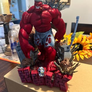 Może przedstawiać: Czerwona figurka akcji muskularnej postaci z siwymi włosami i podartymi niebieskimi szortami. Figurka stoi na podstawie z napisem "HULK" w czerwonych ceglanych literach. Figurka trzyma cygaro i jest otoczona sceną cmentarza.