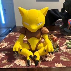 Könnte beinhalten: Eine gelb-braune Actionfigur einer Kreatur mit spitzen Ohren und scharfen Krallen. Die Figur sitzt mit gekreuzten Beinen. Sie hat eine braune Rüstung auf Schultern und Brust. Die Figur steht auf einer Tischdecke mit floralem Muster.