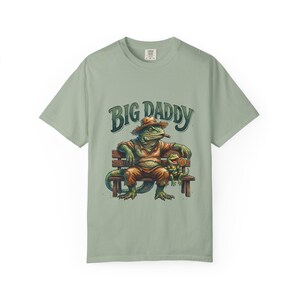 May include: Sage green t-shirt with a graphic of a lizard wearing a hat and smoking a cigar, sitting on a bench with a smaller lizard. The text "BIG DADDY" is arched above the lizards.