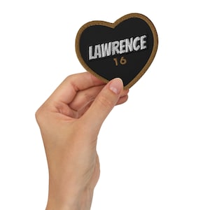 May include: A black heart-shaped patch with a brown border. The patch has white embroidery that reads "LAWRENCE 16".