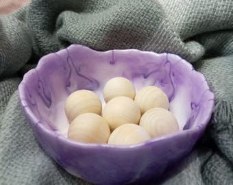 Könnte beinhalten: Eine violett-weiße marmorierte Schale gefüllt mit sechs Holzperlen. Die Schale hat einen welligen Rand und steht auf einem weichen, grauen Stoff.