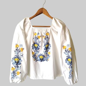 May include: White blouse with long sleeves and a square neckline. The blouse features a floral design in blue and yellow, with the flowers arranged on the front and sleeves. The garment is hanging on a wooden hanger.