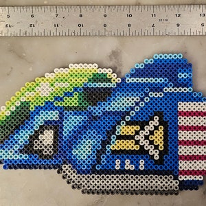 May include: A pixel art design of a blue and white vehicle, possibly a race car or spaceship, made from colorful beads. The design includes details in green, yellow, and red. A ruler is placed above the pixel art.