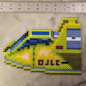 May include: A pixel art creation of a yellow and blue cartoon character, possibly a vehicle or creature. The design is made with small, colorful beads. The piece is approximately 5 inches long, and 2 inches wide.