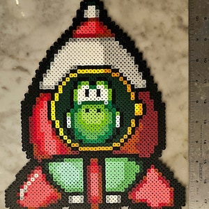 May include: A pixel art creation of a character, likely from a video game, made with colorful plastic beads. The character has a green body, a red hat, and a white brim. The design is outlined in black and has a retro aesthetic.