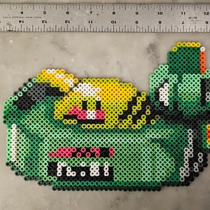 May include: A pixel art creation of a green creature with yellow, black, white, pink, and orange accents. The creature has a large mouth and a smiling expression. The piece is made of small, colorful beads.
