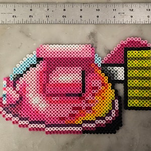 May include: A pixel art design of a pink car with yellow and black accents, created with small, colorful beads. The car has a light blue outline and a black rectangular shape on the side. The design is approximately 5 inches long.
