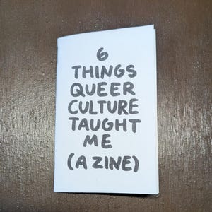 May include: A white zine with a folded spine featuring the black hand-lettered text 6 THINGS QUEER CULTURE TAUGHT ME (A ZINE) centered on the front cover. The booklet is rectangular with a clean white background and bold, dark lettering.