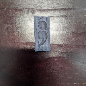 May include: A small, rectangular, blue fabric patch with a semicolon design in a darker shade of blue. The semicolon is centered on the patch, which appears to be made of a textured material. The patch is placed on a dark wooden surface.