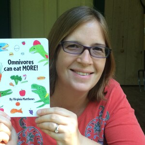 May include: A woman in a red shirt holds up a book titled "Omnivores can eat MORE!" by Virginia Matthews. The book cover features colorful illustrations of dinosaurs and various foods.