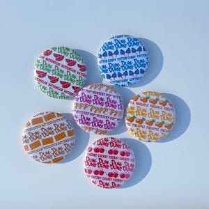 Puede incluir: Cinco botones redondos con diferentes diseños. Cada botón presenta un fondo blanco con un patrón repetido de una fruta o un dulce diferente. Los botones representan sandía, piña, algodón de azúcar, cereza y caramelo de mantequilla.